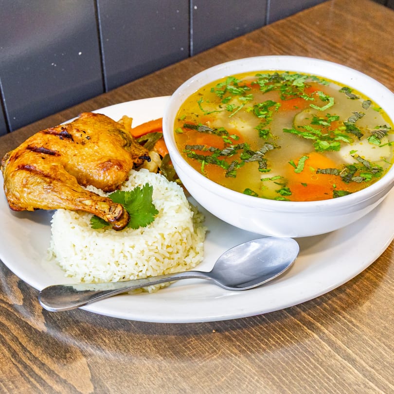 Guatemalan grilled chicken leg and thigh with rice, vegetables, and a bowl of soup.